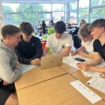 5 students participating in The Richard Huish College Dragons Den 2024. They are sat around a table looking at a laptop.