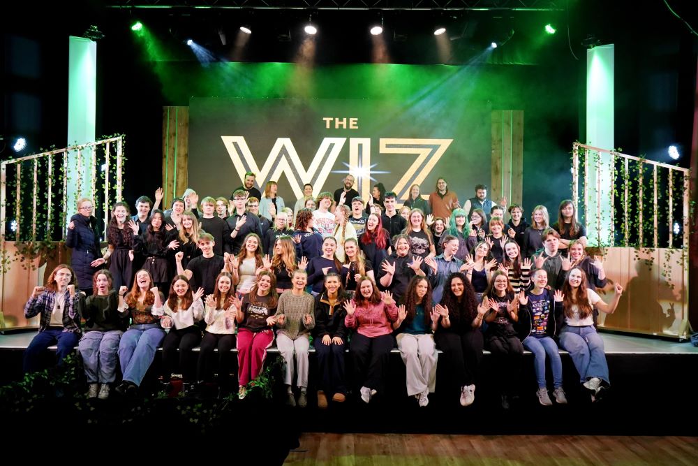 A picture of the entire Wiz cast sat on the stage, with the set around them and green and white lighting. All stage members are smiling and some have their hands in the air.