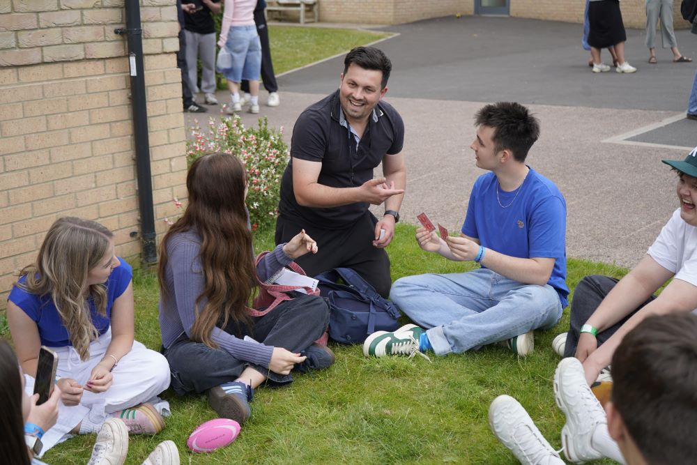 A staff performing card magic tricks with a group of students sat around.
