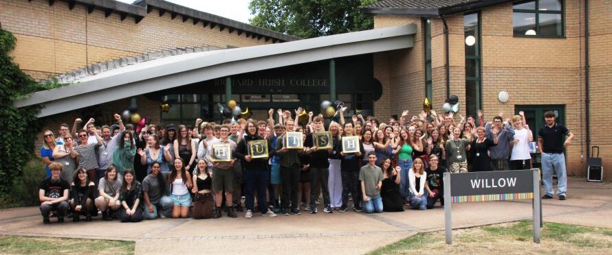 Lots of students stood outside the Willow building, smiling and cheering. Some hold boxes with letters that spell out 'HUISH', There are some balloons too.