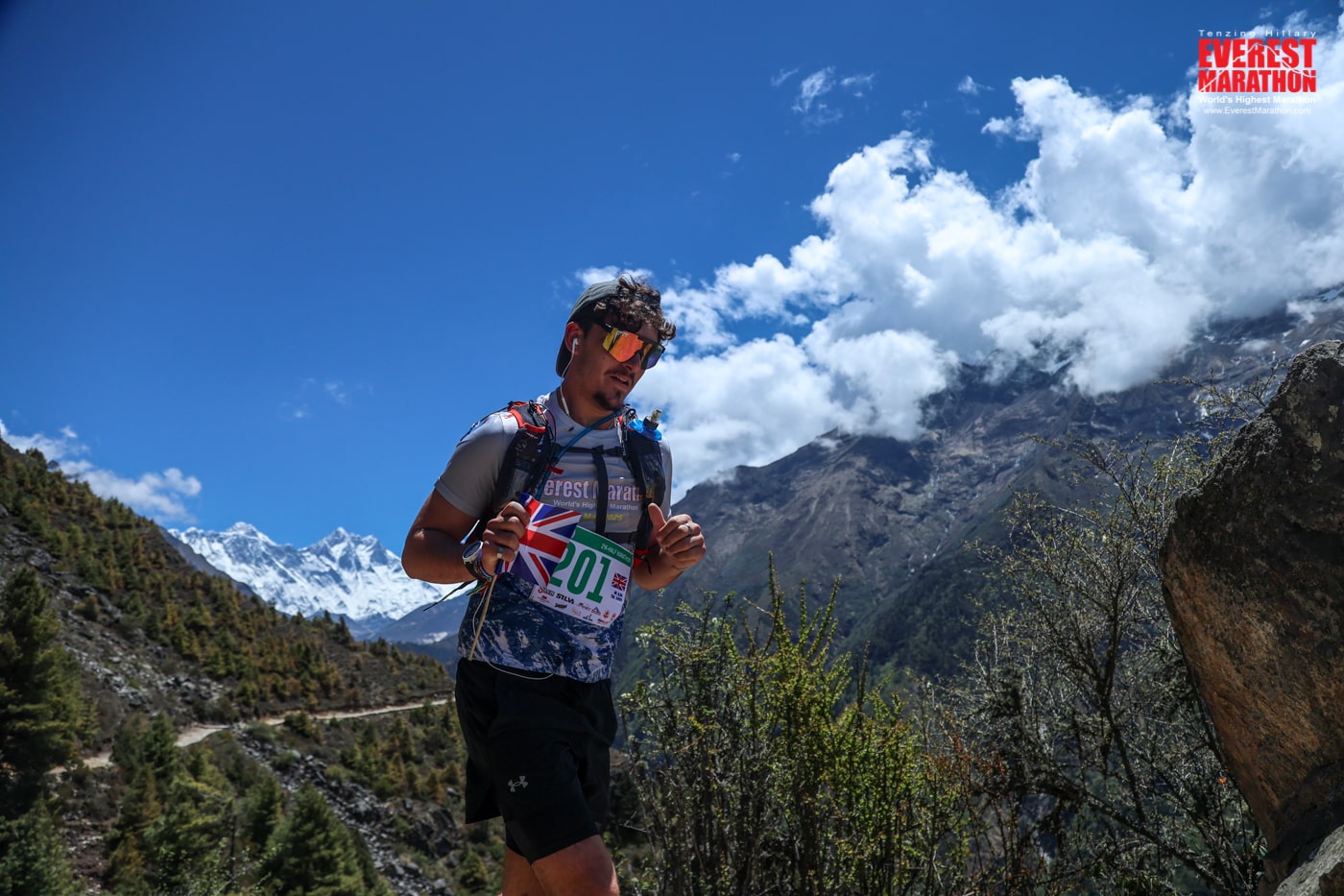 An action shot of Albie running. He is holding a small UK flag, and wears his race number. Clouds and mountain peaks are in the background.