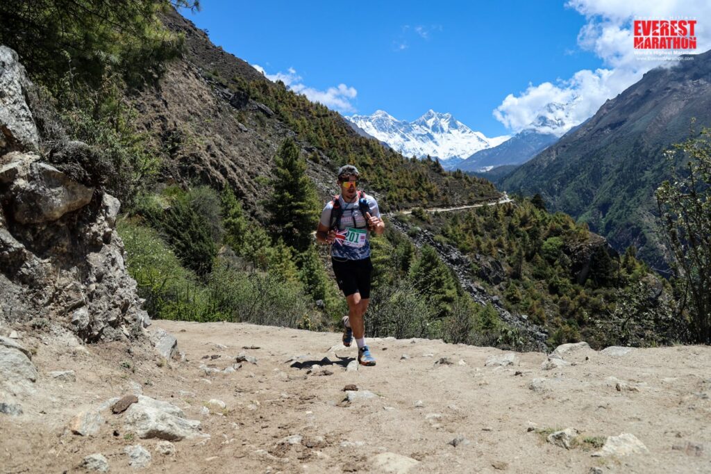 An action shot of Albie running. He is holding a small UK flag, and wears his race number. Clouds, grassy terrain and mountain peaks are in the background.