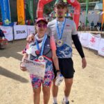 Albie and his mum at the end of the race, each holding a certificate and wearing a medal. The finish line and banners can be seen in the background.