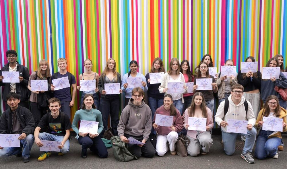 Group of students holding certificates and smiling, standing in front of a vibrant multicoloured striped background.