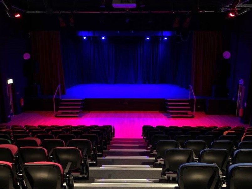 Theatre chairs in the foreground, in front of a stage with purple and pink lighting.
