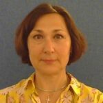 A lady with short brown hair, wearing a yellow shirt and necklace.
