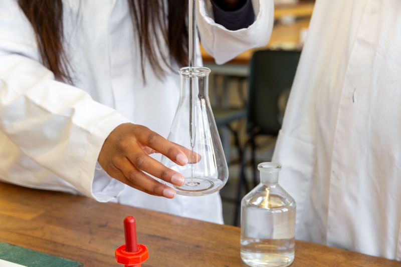 A student in a lab coat holds up a chemistry flask, She is using a pipette to pour liquid into it.