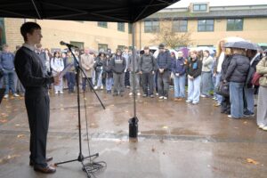A student gives a speech with a microphone, under a gazebo, with students and staff looking on.