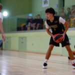 Player in a black and yellow jersey dribbling a basketball while being guarded by an opponent in a red vest, in an indoor basketball court with spectators in the background.
