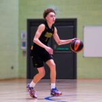 A player in a basketball uniform dribbling a ball across the court during a game in an indoor gymnasium.