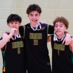Three basketball players in Huish jerseys, numbered 8, 10, and 1, smiling and posing with medals around their necks (biting the medals) in a gym.