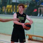 A basketball player wearing a black HUISH jersey numbered 4 passes the ball in an indoor court, facing an opponent in an orange jersey. Spectators are visible in the background.