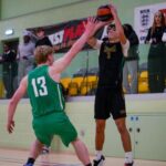 Two basketball players in action on the court, one in a green jersey marked "13" and the other in a black jersey marked "Huish" attempting to catch a basketball. Spectators can be seen in the background.