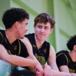 Three basketball players in black and yellow uniforms sitting on the sidelines, engaging in a conversation during a break in the game.