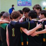 A basketball team in green and gold uniforms huddling together on the court.