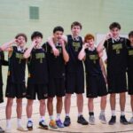 A group of nine basketball players in black uniforms with Huish written on the front, joyfully posing with their mdeals (they are biting them).