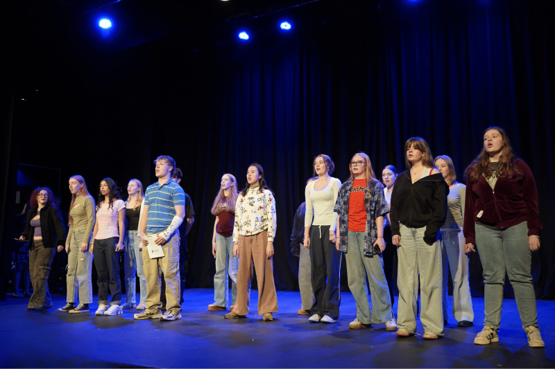 Group of student actors on the stage during a Dear Evan Hansen rehearsal in the Huish Theatre. There is blue lighting on the stage.