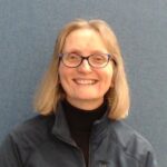 A lady with short hair and glasses, and a black polo neck top and blue fleece, smiling at the camera.