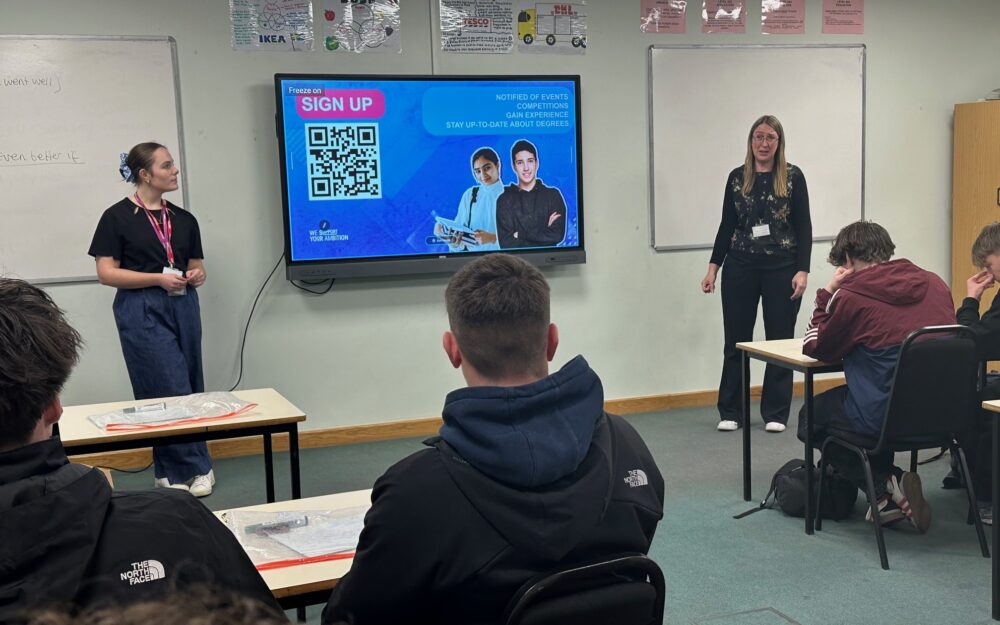 Two educators stand in a classroom presenting to students seated at tables. A large screen displays a "SIGN UP" message with a QR code and an image of a person. Various educational posters are visible on the walls.