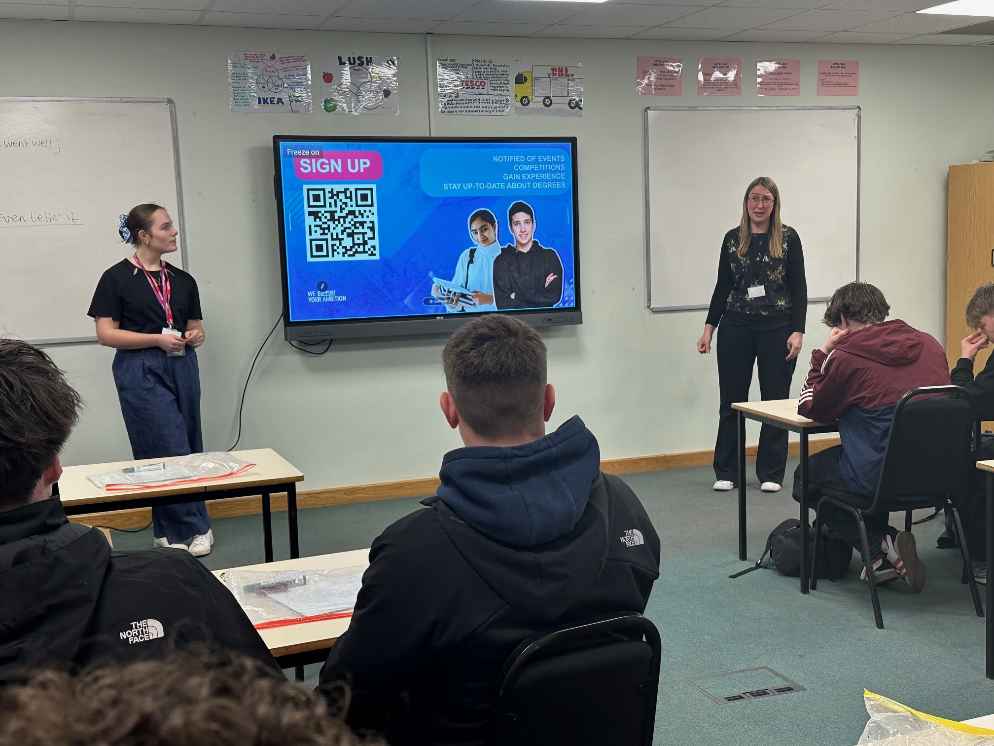 Two educators stand in a classroom presenting to students seated at tables. A large screen displays a "SIGN UP" message with a QR code and an image of a person. Various educational posters are visible on the walls.