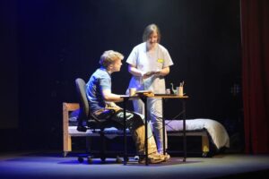 A performer sits in a chair beside a bed while another performer in hospital scrubs stands nearby, holding papers.