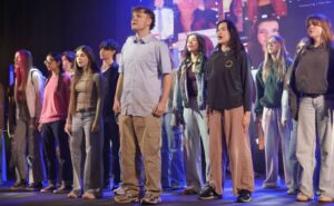 A large group of performers stands onstage in casual clothing, arranged in rows, with bright stage lighting and a colourful digital backdrop behind them.