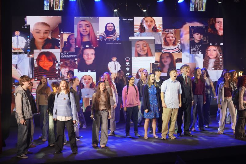 A group of performers stands on stage, with large digital screens behind them displaying a collage of social media-style videos and images. The background and lighting suggest a lively and dynamic atmosphere.