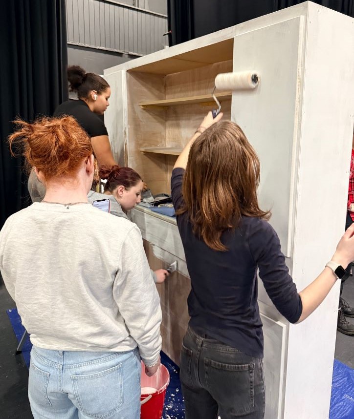 A group of stage crafts students working together to paint a large set piece with shelving using paint rollers.
