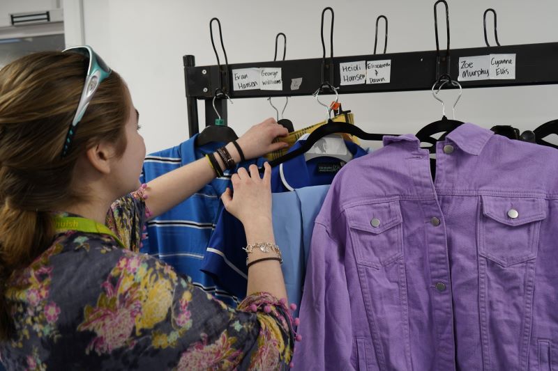 A person is browsing a clothing rack with various outfits. Labels on the rack have the character and actors' names. The individual is adjusting a blue garment that is hung among other colourful clothes.