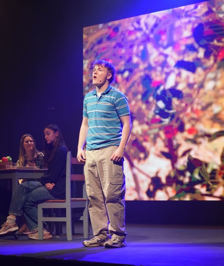 A person stands on stage performing in front of a colourful screen showing leaves. Two people sit at a table in the background, and the scene is lit with blue stage lighting.