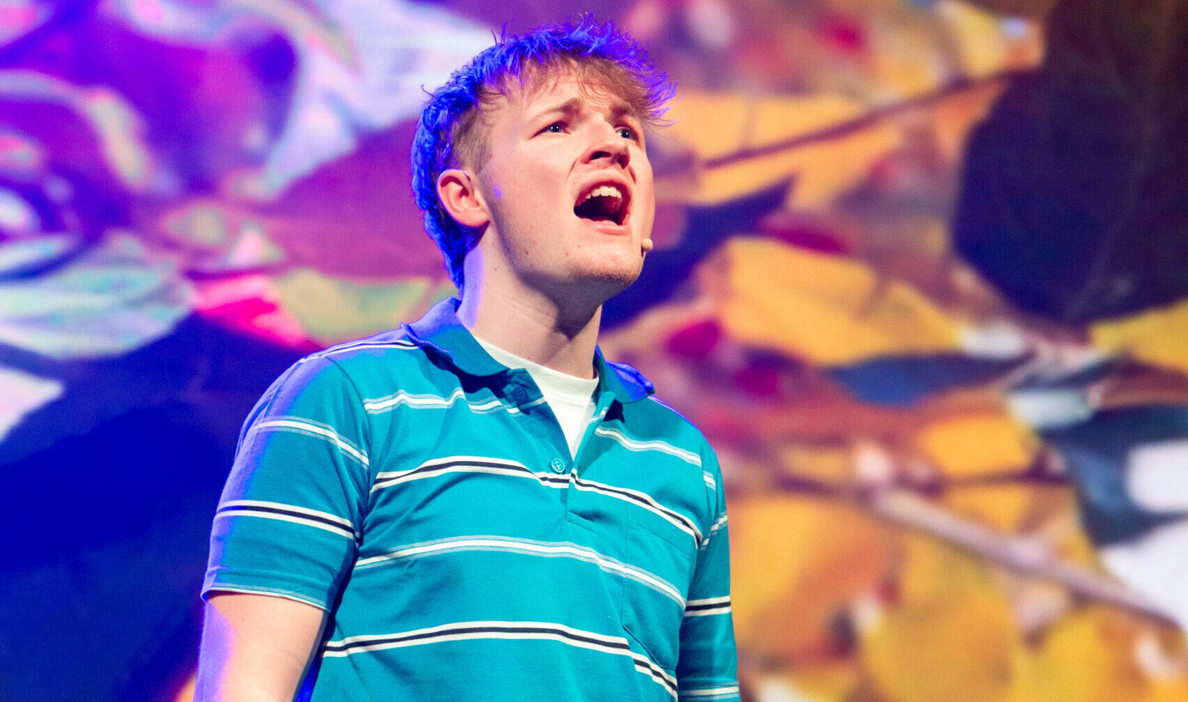 A boy sings on stage in front of a digital leaf backdrop. He has a microphone on his face and wears a stripy blue top and cargo trousers. One of his arms is in cast.
