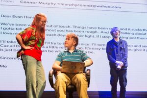 Three performers stand in front of a large projected email on a screen while one sits with a laptop covered in stickers.