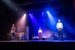 Three performers stand spaced across a stage set that includes a kitchen area and a bedroom, each lit by separate spotlights.