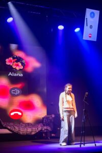 A person stands on a stage in front of a bedroom set-up with bed and duvet. She is facing a camera on a tripod. The background features bright, abstract digital icons including a speaker and a phone symbol, which makes it look like a phone call is taking place.