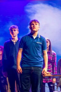 A performer stands at the front of the stage in a blue polo shirt, with other cast members standing behind against a sky-themed background.
