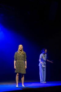 Two performers stand apart onstage, one wearing a patterned dress and the other in medical scrubs, under dramatic blue lighting suggesting a tense or emotional moment.