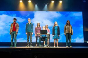 A group of six performers stands in a line onstage. Another performer is sat at a desk with a laptop in front of him. The backdrop is a large sky with fluffy white clouds.
