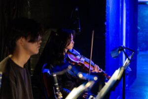 Musicians sit in the orchestra area beside the stage, including a violinist illuminated by blue stage light, with music stands and microphones around them.