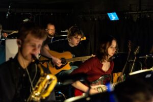 Members of the orchestra play instruments such as acoustic and electric guitars, seated in a dimly lit pit area beneath the stage.
