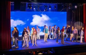 Performers walk and stand across a wide stage with a bright sky backdrop, creating a dynamic ensemble moment.