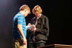 Two performers stand close together onstage as one writes on the other’s arm cast.