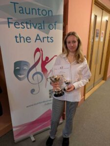 A student holds two trophies and a certificate whilst stood in front of a banner that reads 'taunton festival of the arts'.