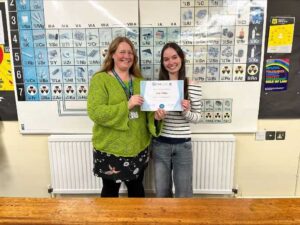 A student holds a certificate whilst stood next to a teacher. They stand in front of a periodic table poster.