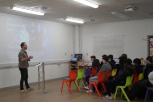A group of people sit in a classroom, paying attention to a speaker standing at a podium. Behind the speaker, a presentation slide is displayed on a whiteboard. The slide includes the text 'The Business of Sports', an image of sports figures, and the name "Rhys Beer." The classroom is equipped with colourful chairs and wood flooring.