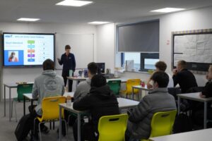 A classroom setting with a person standing at the front presenting to a group of seated individuals. A screen displays a slide titled "Who am I: My Career Pathway" and a name, "Steph Morland." The room is equipped with desks and chairs.