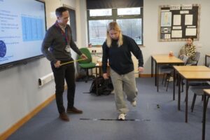 A person is stepping over a black line on the floor, about to do a hop test, in a classroom while another individual stands nearby holding a yellow measuring tape. The wall displays a BTEC Sport sign and there is also a screen with text on it. A person is seated in the background.