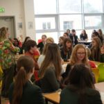 A facilitator speaks to a room that is made up of groups of students sat at tables.