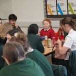 A group of students sit around a table in a classroom, participating in a workshop activity. Drinks, papers, and stationery are spread across the table. Posters on the wall behind them display information about Early Years Education and Teaching Assistant courses. The setting is a busy, collaborative learning environment.