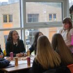 A group of students sits around a table in front of a large window, engaging in a guided discussion. A person in a bright jumper stands beside the table and a younger student, with a school building visible outside.