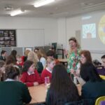 A facilitator speaks to a table of students. A projector displays Citizens UK content on the wall.